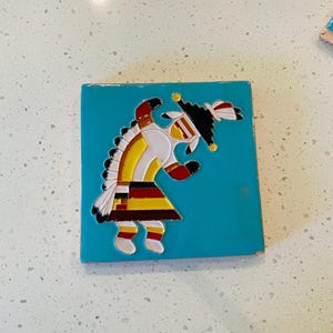 May include: A square, turquoise tile featuring a Native American figure in white, yellow, red, and black. The figure is depicted in a stylized, dancing pose. The tile is likely ceramic or a similar material.