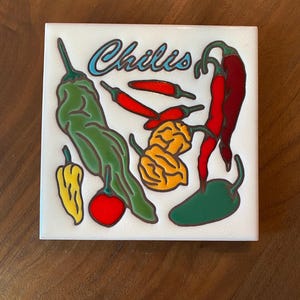 May include: Square white tile featuring colorful illustrations of various chili peppers. The word "Chilis" is written in a blue, cursive font. The peppers are green, red, yellow, and orange, with green stems.