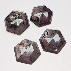 May include: Four faceted, hexagonal gemstones with a dark purple hue and internal inclusions. The gems are arranged on a white surface, showcasing their geometric shapes and reflective surfaces. The stones are likely for jewelry making or collecting.