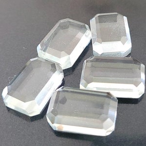 May include: Five clear, faceted, rectangular gemstones. The gemstones are arranged in a cluster on a grey surface.