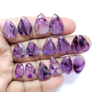 Puede incluir: Una colección de 13 cabujones de piedra preciosa de amatista púrpura en forma de lágrima. Las piedras son translúcidas y tienen una variedad de tonos púrpura.