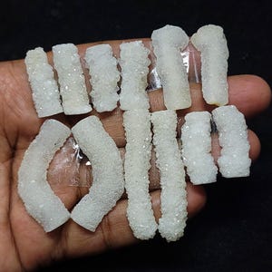 May include: A collection of small, white, crystal-covered mineral specimens. The crystals have a rough, textured appearance and are arranged in various shapes, including curved and cylindrical forms. The crystals are held in a hand against a dark background.