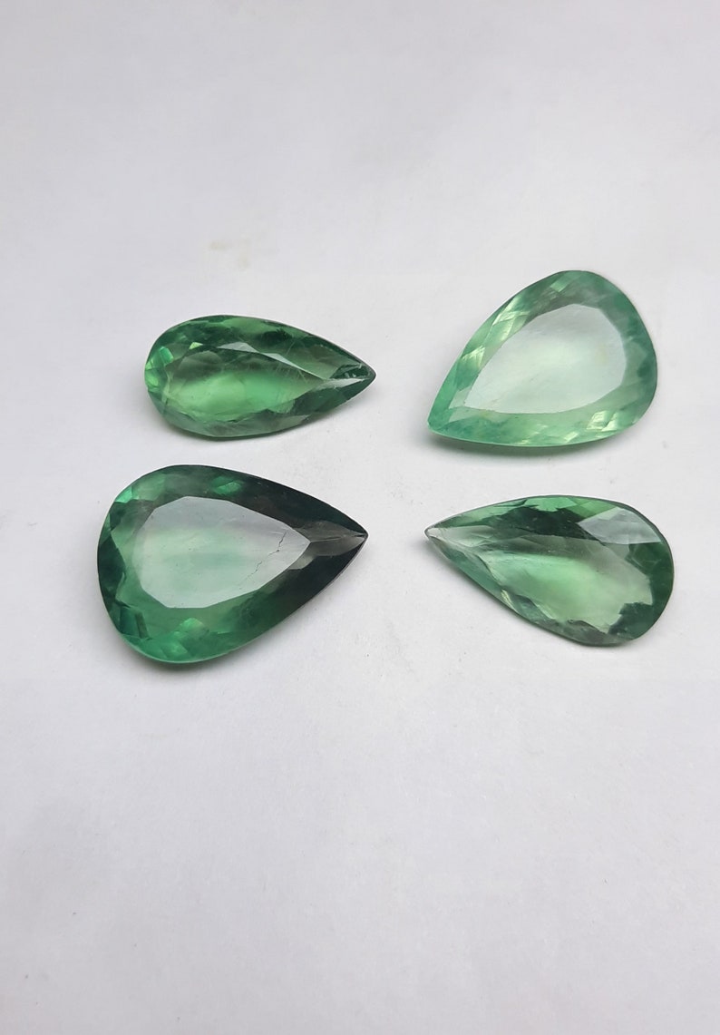 May include: Four pear-shaped green gemstones on a white background. The gemstones are faceted and have a translucent appearance.