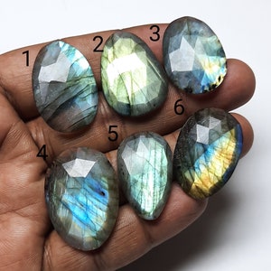May include: Six faceted labradorite gemstones in various shapes and sizes. The stones display a range of colors, including blues, greens, and yellows, with iridescent flashes. Each stone is held in a hand, showcasing their polished surfaces and unique patterns.