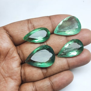 May include: Four pear-shaped green gemstones, likely emeralds, are displayed on a hand. The gemstones are faceted and have a translucent appearance.