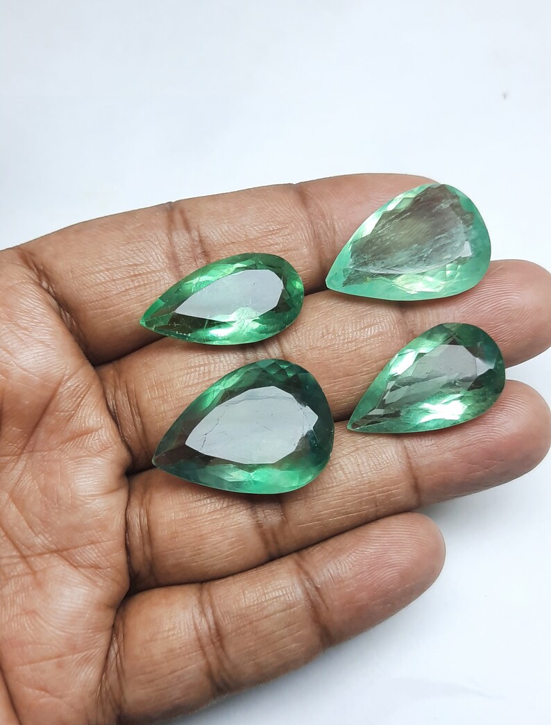 May include: Four pear-shaped green gemstones, likely emeralds, are displayed on a hand. The gemstones are faceted and have a translucent appearance.