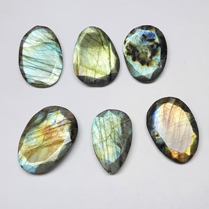 May include: Six polished labradorite gemstones in various shapes, including oval and teardrop. The stones display iridescent flashes of blue, green, and gold, with a faceted cut. The gemstones are set against a white background.