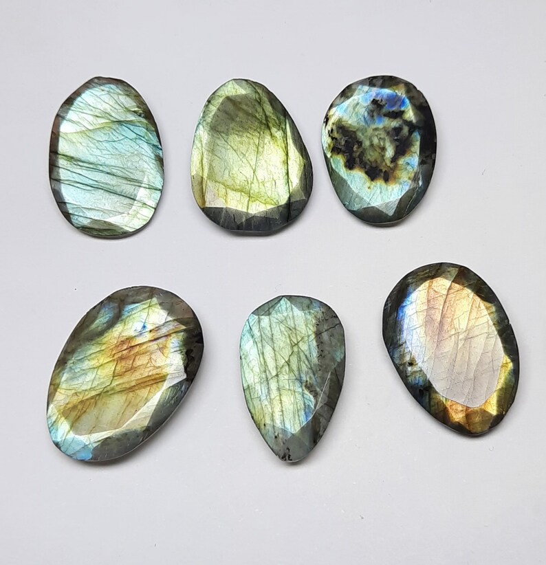 May include: Six polished labradorite gemstones in various shapes, including oval and teardrop. The stones display iridescent flashes of blue, green, and gold, with a faceted cut. The gemstones are set against a white background.