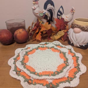 Puede incluir: Dos tapetes redondos tejidos a mano al ganchillo con un borde festoneado. El diseño consiste en un círculo central de color crema rodeado por un anillo de motivos de calabazas naranjas y un borde fino verde salvia, acabado en crema.