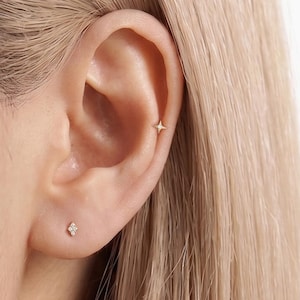 May include: Two small gold earrings are worn in the ear. One earring is a four-pointed star shape located in the upper cartilage, and the other is a small cluster of four tiny stones positioned in the earlobe, both featuring a metallic finish.