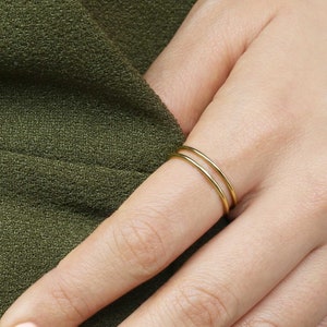 Puede incluir: Primer plano de una mano con un fino anillo de doble banda de oro. El anillo está en el dedo anular de la mano derecha. La mano está apoyada sobre una superficie de tela verde.