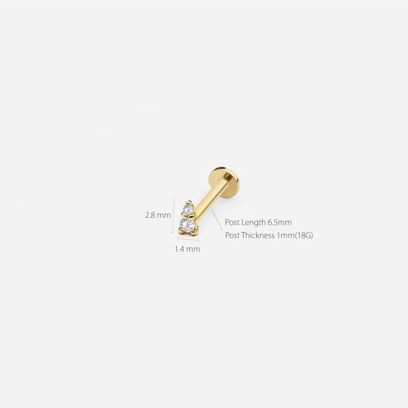 May include: Gold body jewelry with two clear gemstones. The jewelry is 2.8 mm wide and 1.4 mm thick. The post is 6.5 mm long and 1 mm thick (18 gauge).
