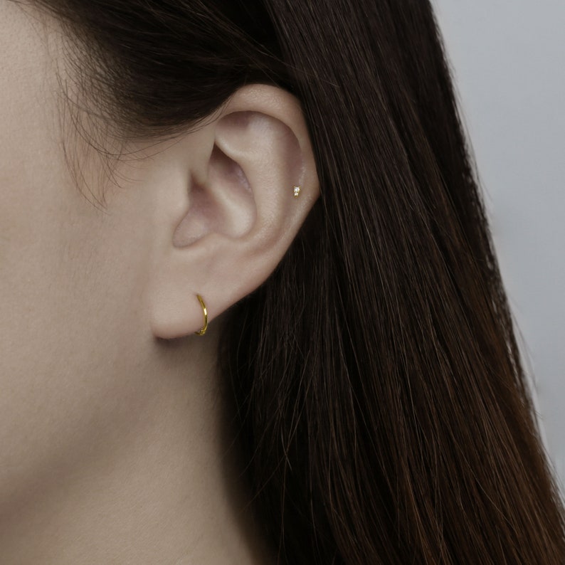 May include: A close-up of a person's ear with a small gold hoop earring in the lobe and a tiny gold stud earring in the cartilage.