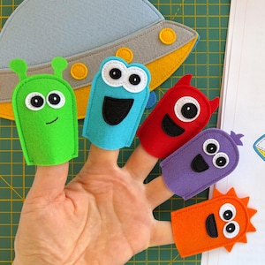 Op de afbeelding: Een hand met vijf kleurrijke vingerpoppetjes van vilt die vriendelijke ruimtemonsters uitbeelden. De poppetjes zijn groen, blauw, rood, paars en oranje. Een vilt ruimteschip en een naaipatroon getiteld "Little Space Monsters in a Flying Saucer" zijn ook zichtbaar.