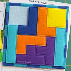 Könnte beinhalten: Ein farbenfrohes Filz-Block-Puzzle-Spiel mit dem Text "Block Build Puzzle Game". Das Spiel enthält verschieden geformte Blöcke in Rot, Grün, Blau, Gelb, Orange und Lila. Das Spiel befindet sich auf einem grünen Gitterhintergrund.