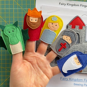Könnte beinhalten: Eine Hand zeigt fünf Fingerpuppen aus Filz: einen grünen Drachen, einen König, eine Prinzessin, einen Ritter und einen Zauberer. Eine graue Burg aus Filz und ein Papier mit dem Text "Fairy Kingdom Finger Puppets" befinden sich im Hintergrund. Die Puppen sind farbenfroh und detailliert.