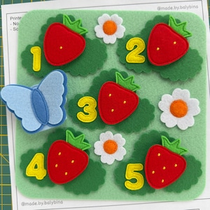 Könnte beinhalten: Ein hellgrünes Filz-Aktivitätsbrett mit fünf roten Erdbeeren mit grünen Blättern, fünf gelben Zahlen von eins bis fünf, drei weißen Blumen mit orangefarbenen Zentren und einem hellblauen Schmetterling, alles auf einem grünen Hintergrund.