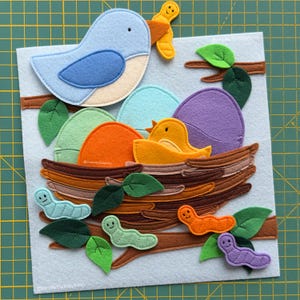 May include: A felt craft set featuring a light blue bird holding a yellow worm, a brown nest with colourful eggs, a small yellow chick, and several green, orange, and purple worms on brown branches with green leaves on a light grey felt base.