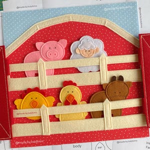 Op de afbeelding: Een rood vilt knutselproject in de vorm van een schuur met witte hekwerk latten en vijf vilten dierenfiguren, waaronder een roze varken, een wit schaap, een gele kip, een geel kuiken en een bruin paard, met twee rode vilten deuren aan de zijkanten.