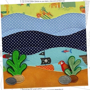 Könnte beinhalten: Ein Filz-Applikations-Projekt für eine Kinderspielmatte. Das Bild zeigt ein blaues und weißes wellenförmiges Meer, einen dunkelblauen und weiß gepunkteten Meeresboden, einen grün-orangenen Strand und ein Piratenschiff mit einer Totenkopf- und Knochenflagge. Es gibt auch zwei grüne Seetangpflanzen und zwei graue Steine.