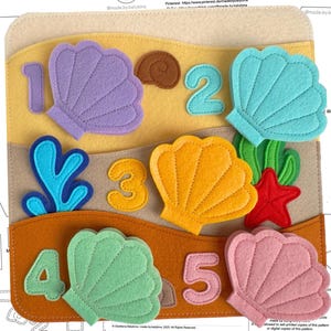 Könnte beinhalten: Ein pädagogisches Spielzeug aus Filz mit dem Titel "Seashell Hunt" mit bunten Filzmuscheln, Zahlen und Meereselementen. Das Spielzeug hat einen hellbeigen Hintergrund mit gelben, braunen und orangen Abschnitten. Die Zahlen 1-5 werden mit verschiedenfarbigen Muscheln angezeigt.