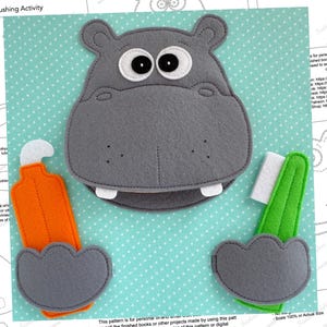 Könnte beinhalten: Ein Filz-Bastelmuster für ein Hippopotamus mit einem Mund, der sich öffnet, um Zähne zu zeigen. Das Muster enthält eine Zahnbürste, Zahnpasta und einen Hippopotamus-Kopf. Das Muster ist auf einem hellblauen Hintergrund mit weißen Punkten gedruckt. Hippopotamus-Zähneputzaktivität.