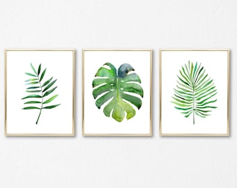 May include: Three framed watercolor paintings of tropical leaves. The paintings are in gold frames and feature a green palm leaf, a green monstera leaf, and a green fern leaf.
