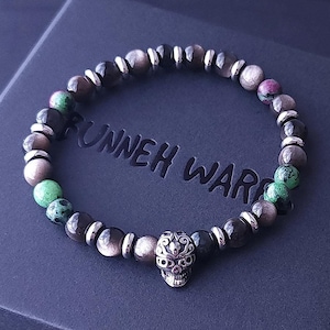 May include: A black box with a red ribbon on the front. Inside the box is a bracelet made of black, green, and brown beads with a silver skull charm. The box has the text "UNNEH WAR" printed on the top.