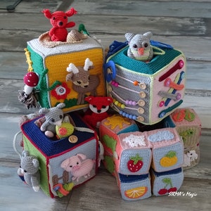 May include: A stack of six colourful crocheted blocks with animal and nature-themed designs. The blocks are made of soft yarn and feature a variety of textures. The top block has a red fox, the second block has a green owl, the third block has a deer, the fourth block has a rabbit, the fifth block has a pig, and the sixth block has a strawberry, lemon, and cherry.