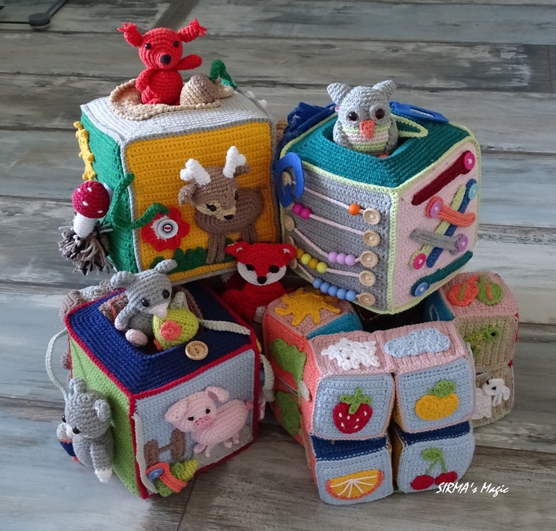 May include: A stack of six colourful crocheted blocks with animal and nature-themed designs. The blocks are made of soft yarn and feature a variety of textures. The top block has a red fox, the second block has a green owl, the third block has a deer, the fourth block has a rabbit, the fifth block has a pig, and the sixth block has a strawberry, lemon, and cherry.