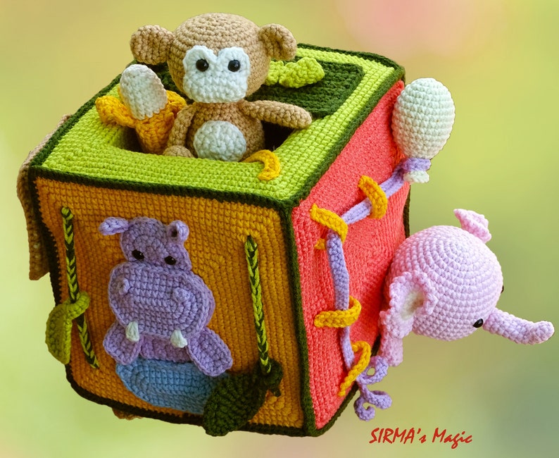 May include: A handmade crochet cube toy featuring a monkey with a banana, a hippo, and an elephant. The cube is multi-colored with green, orange, yellow, and pink sections. The toy has a whimsical design, perfect for children.