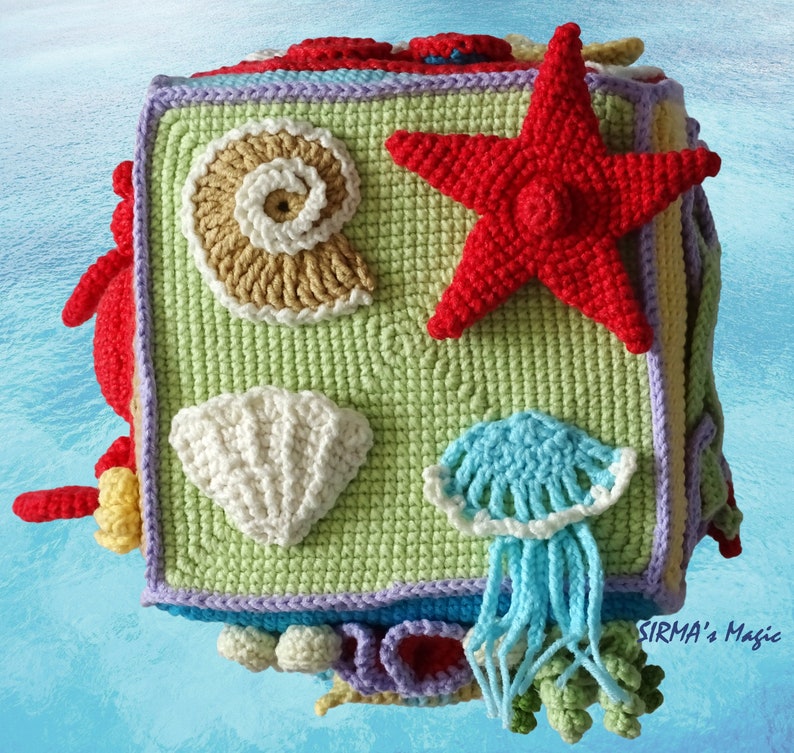 May include: A crocheted cube toy featuring a variety of sea creatures, such as a starfish, seashell, and jellyfish. The cube is crafted from different coloured yarn and has a textured finish. The text "SIRMA's Magic" is visible in the bottom right corner.