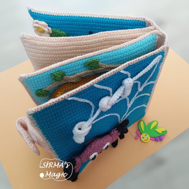May include: A handmade crochet book with blue, beige, and white accents. The pages feature various designs, including a flower, a branch with leaves, and a spiderweb. A purple and black creature and a butterfly are also present. The book is labeled "SIRMA's Magic".