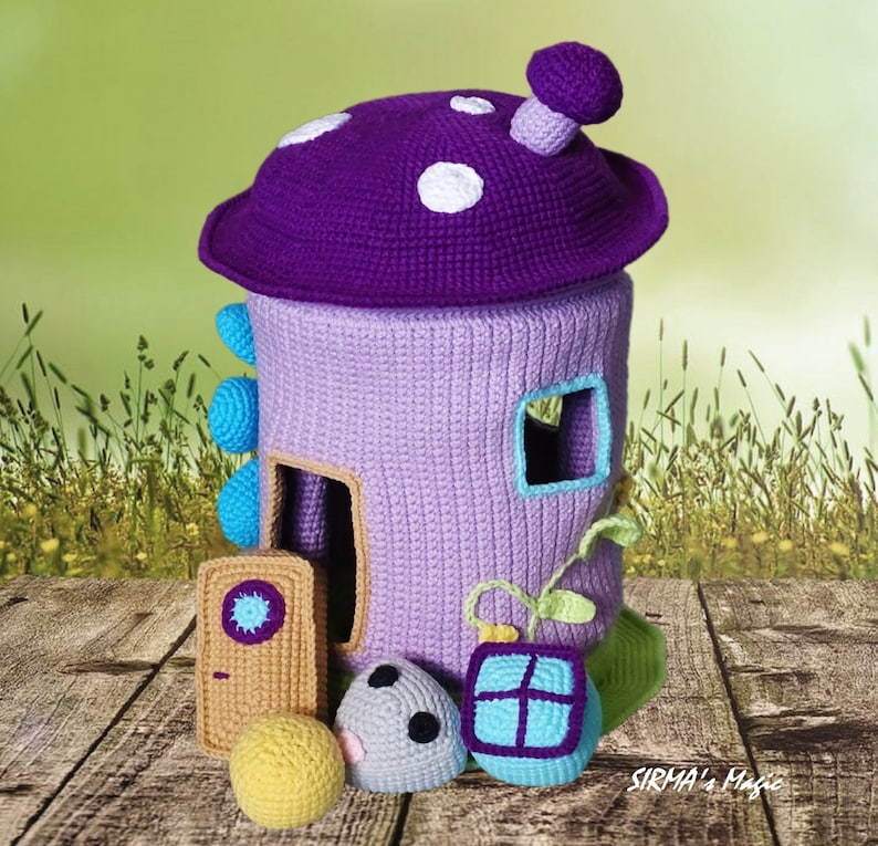 May include: A crocheted purple mushroom house with white polka dots, a blue door, and a window. The house is surrounded by green grass and a wooden floor. There are also three crocheted balls in front of the house.