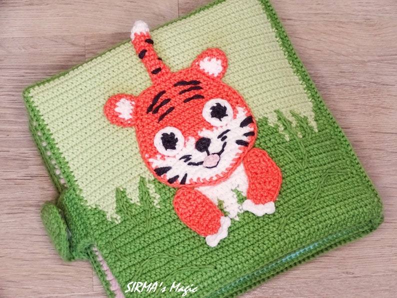 May include: A green crocheted baby book with a cute orange and white tiger applique. The book has a flap on the side and the text "SIRMA's Magic" is visible on the bottom.