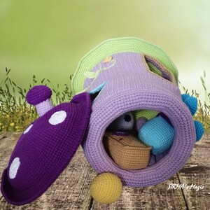 May include: A crocheted purple and green playhouse with a removable lid and a variety of colorful crocheted balls inside. The playhouse is sitting on a wooden surface with a green grassy background. The text "SIRMA's Magic" is visible on the bottom right corner of the image.