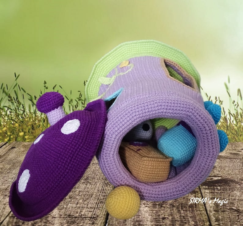 May include: A crocheted purple and green playhouse with a removable lid and a variety of colorful crocheted balls inside. The playhouse is sitting on a wooden surface with a green grassy background. The text "SIRMA's Magic" is visible on the bottom right corner of the image.