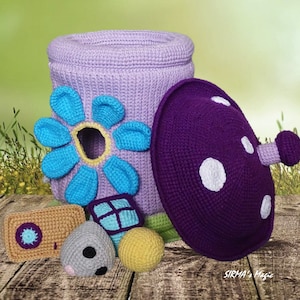 May include: A crocheted flower-shaped toy storage bin with a purple crocheted mushroom lid. The bin is light purple with a blue flower and a yellow center. The mushroom lid has white polka dots. There are four smaller crocheted toys in front of the bin, including a gray mouse, a yellow ball, a blue and green block, and a brown rectangle with a blue circle.