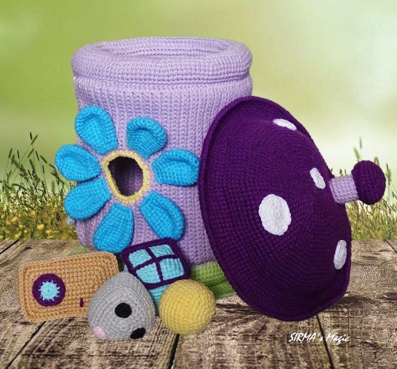 May include: A crocheted flower-shaped toy storage bin with a purple crocheted mushroom lid. The bin is light purple with a blue flower and a yellow center. The mushroom lid has white polka dots. There are four smaller crocheted toys in front of the bin, including a gray mouse, a yellow ball, a blue and green block, and a brown rectangle with a blue circle.