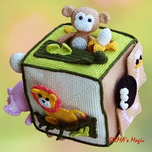 May include: A handmade crochet cube toy with a jungle theme. The cube features a monkey, banana, and lion, with green and brown accents. The cube is beige and green, with a pink flower and a white ball on the sides.