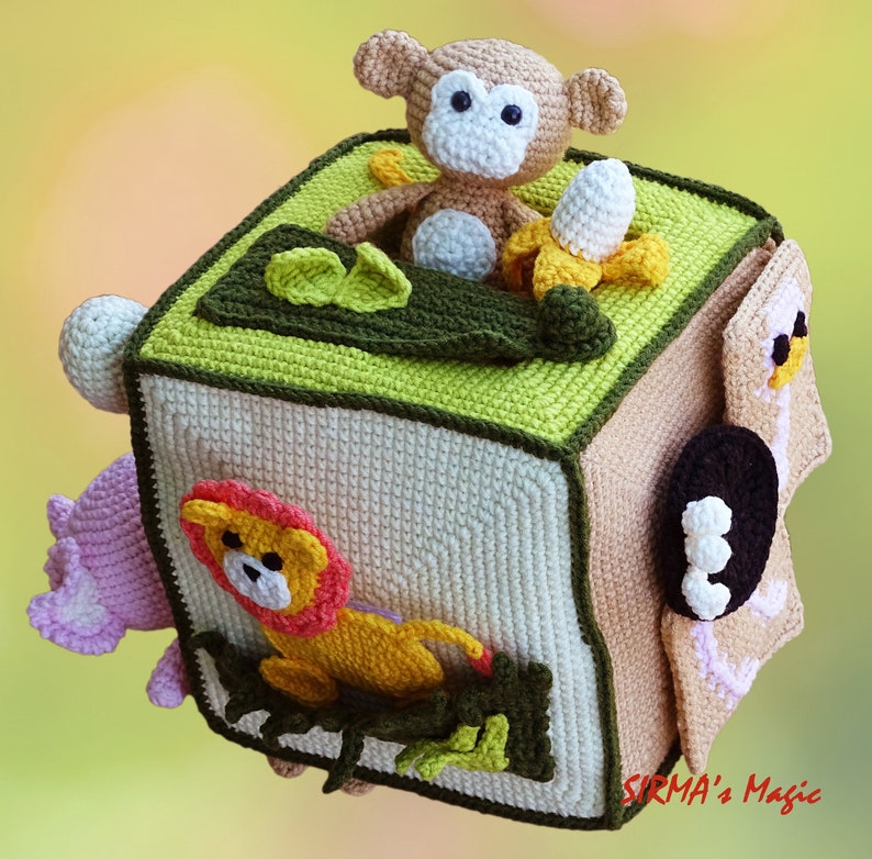 May include: A handmade crochet cube toy with a jungle theme. The cube features a monkey, banana, and lion, with green and brown accents. The cube is beige and green, with a pink flower and a white ball on the sides.