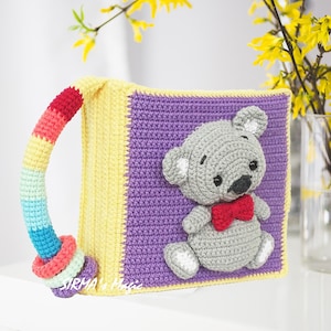May include: A crocheted book with a rainbow handle and a gray crocheted teddy bear with a red bow tie on the front. The book is purple and yellow.