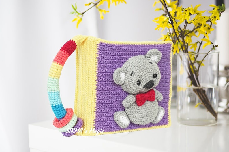 May include: A crocheted book with a rainbow handle and a gray crocheted teddy bear with a red bow tie on the front. The book is purple and yellow.