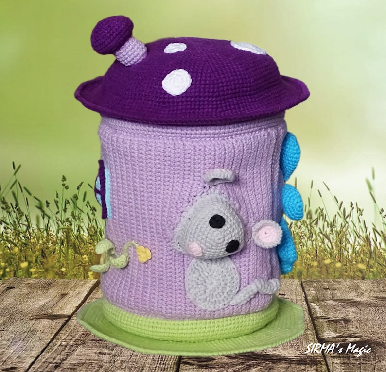 May include: A crocheted mushroom-shaped toy storage bin with a purple top, white polka dots, and a light purple body. The bin features a crocheted gray mouse on the side and a blue flower on the back. The bin is sitting on a wooden surface.