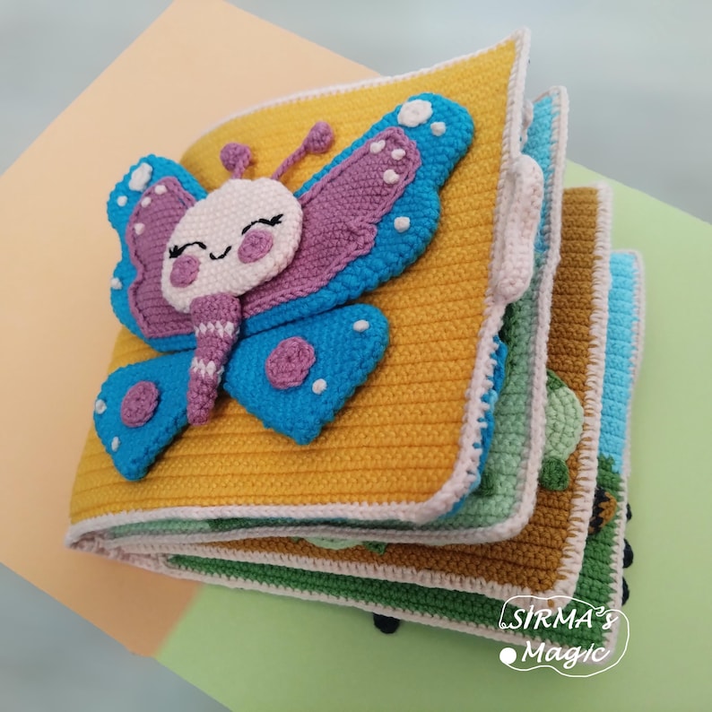 May include: A handmade crochet book with a yellow cover and a large, smiling butterfly applique in blue, purple, and white. The book has multiple colorful pages and the text "SIRMA'S Magic".