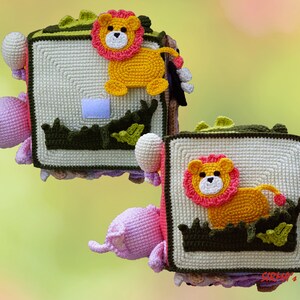 May include: Crocheted soft book featuring a lion design. The lion has a yellow body and a pink mane, set against a cream-colored background. The book has a green border and additional crocheted elements, with a pink flower and a white ball.