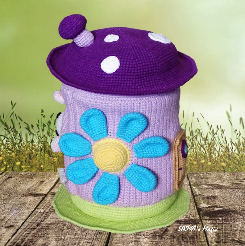May include: A crocheted mushroom-shaped toy with a purple top, white polka dots, and a light purple body. The body has a blue flower with a yellow center and a brown door with a small window.