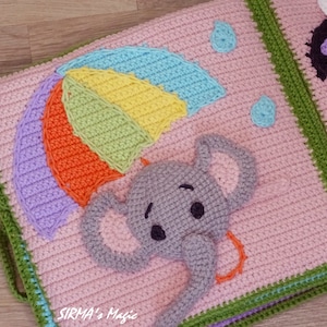 May include: A crocheted baby book page with a pink background, a rainbow umbrella, and a grey elephant with a trunk. The page is bordered with green and blue crochet.
