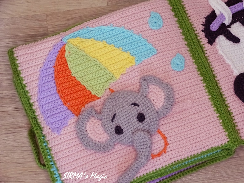 May include: A crocheted baby book page with a pink background, a rainbow umbrella, and a grey elephant with a trunk. The page is bordered with green and blue crochet.
