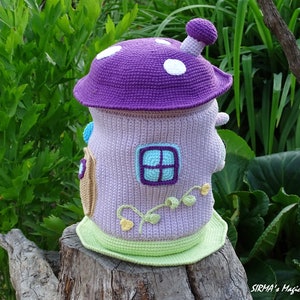 May include: A crocheted mushroom-shaped house with a purple roof, white polka dots, and a green base. The house has a small window with a blue frame and a light purple background. The house is sitting on a tree stump.